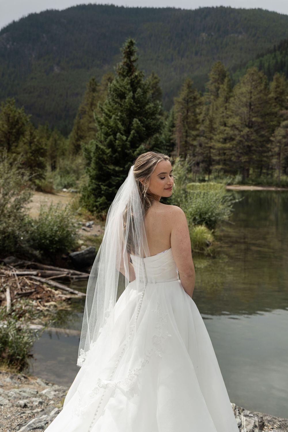 Bride in a strapless white ball gown and veil poses by a mountain lake surrounded by evergreen trees.