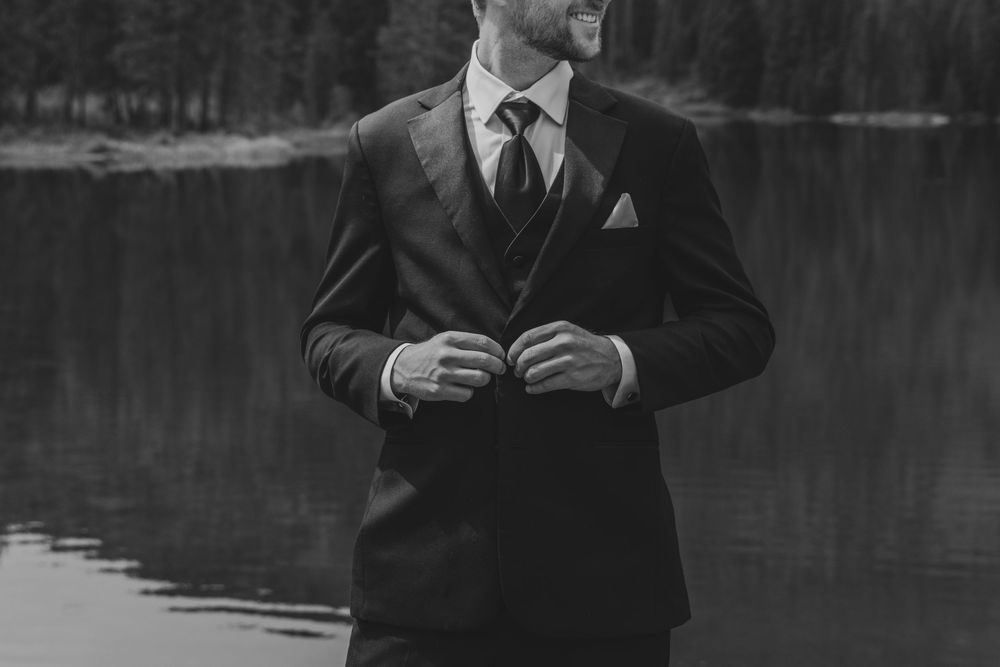 Black and white photo of a groom in a dark suit adjusting his jacket near a serene lake surrounded by trees.
