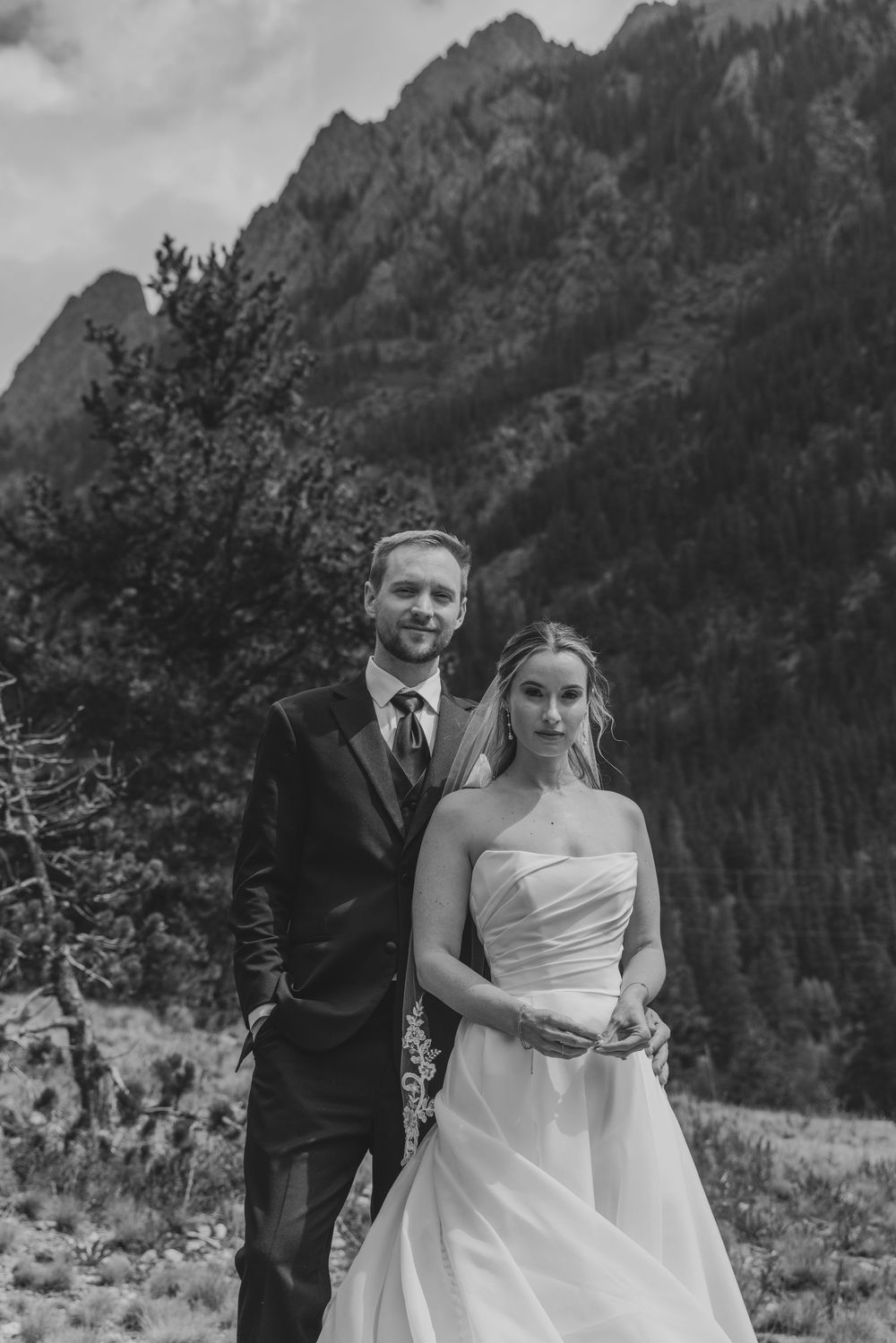 Black and white portrait of smiling bride and groom posing before rugged mountain backdrop.