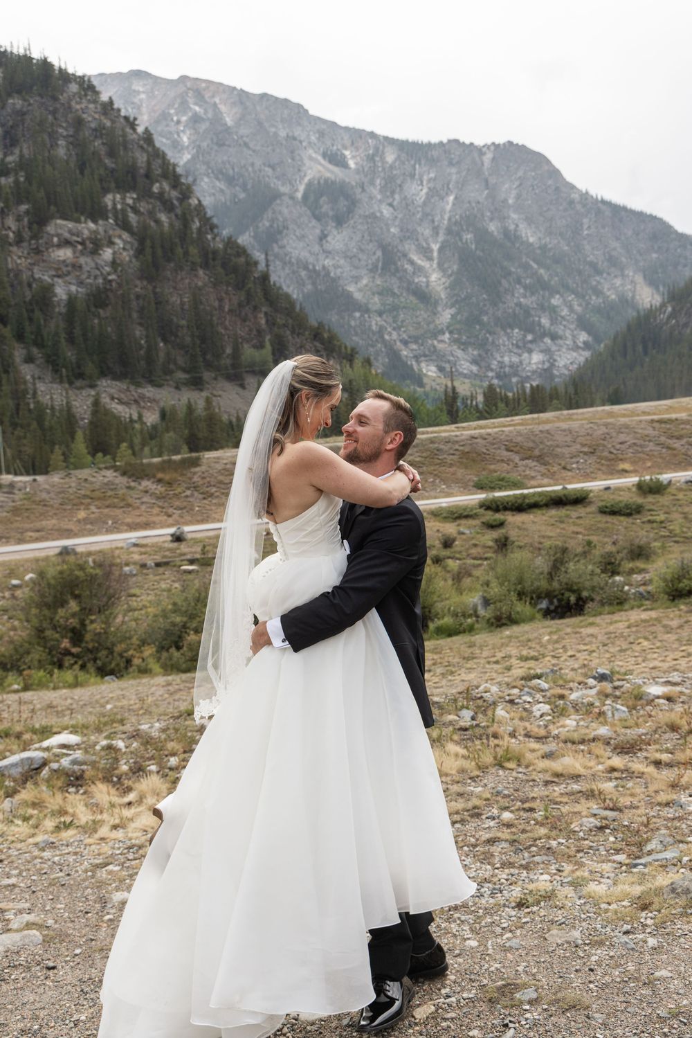 Groom lifting bride in wedding dress with mountain backdrop during outdoor elopement ceremony.