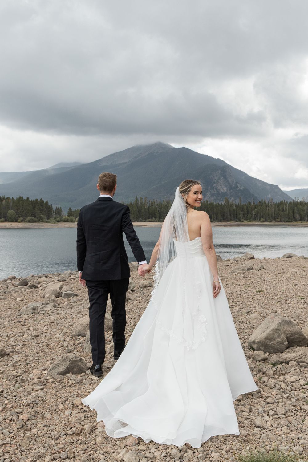 Bride in white gown and veil holds grooms hand by rocky mountain lake under cloudy skies.