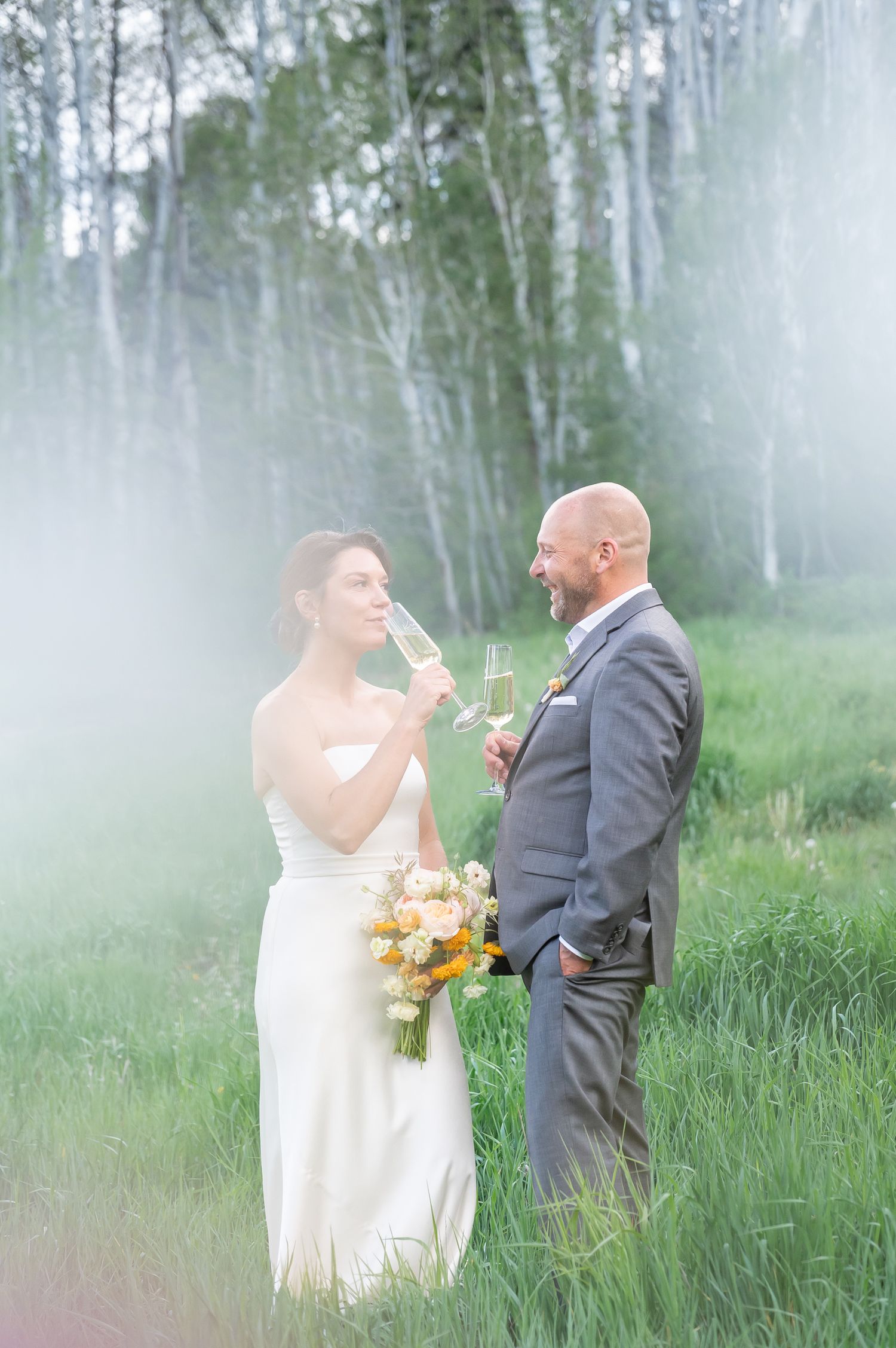 Bride and groom toasting with champagne in a misty forest meadow, bride holding a colorful bouquet in a white gown for Colorado elopement