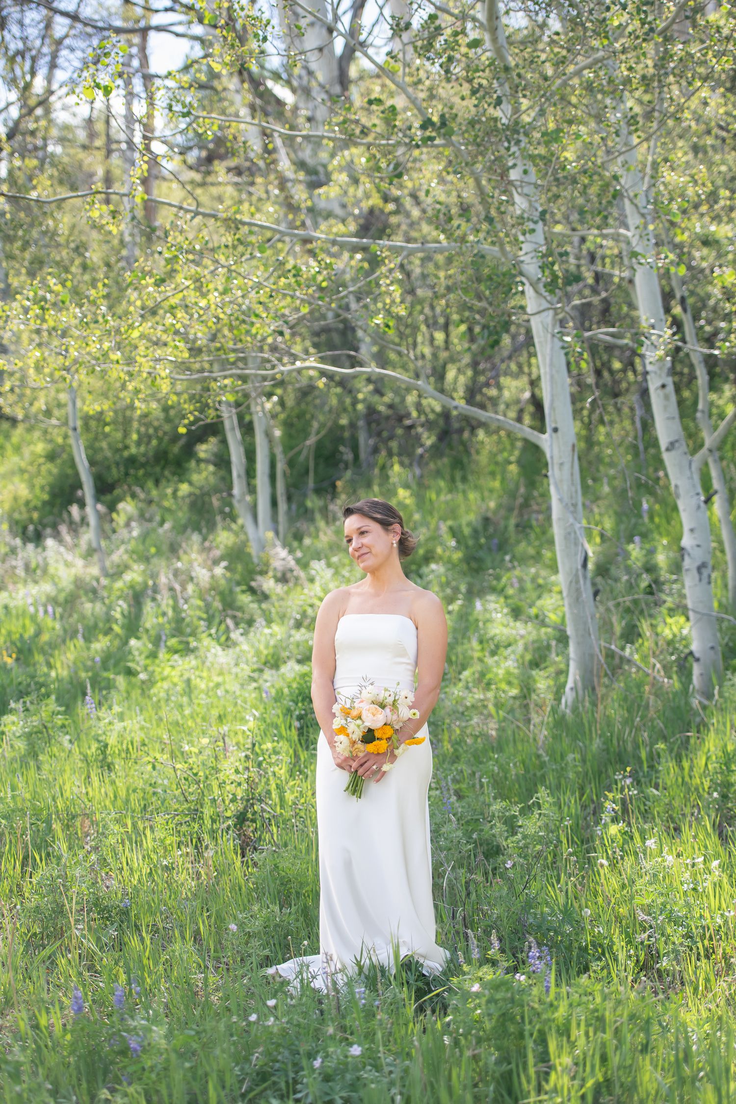 Bride in white strapless gown holding yellow bouquet stands among green aspen trees in a sunlit meadow at a Colorado elopement