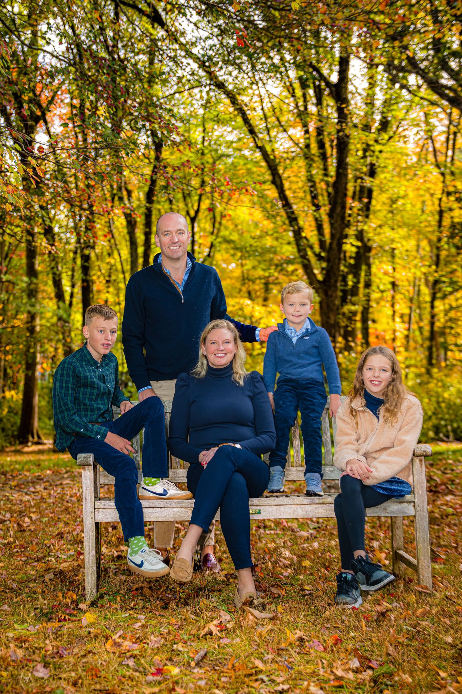 Family of five posing on a wooden bench surrounded by colorful autumn foliage in a scenic outdoor setting.