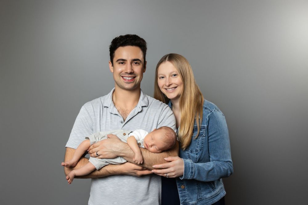 A smiling young couple holding a newborn baby together against a gray studio background.