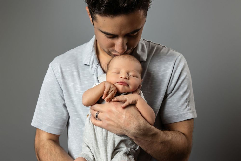 A young father tenderly kisses his sleeping newborn baby while cradling the infant against his chest.