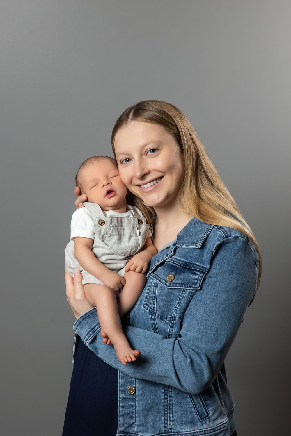 Smiling blonde mother in denim jacket holding her newborn baby against a gray studio background.