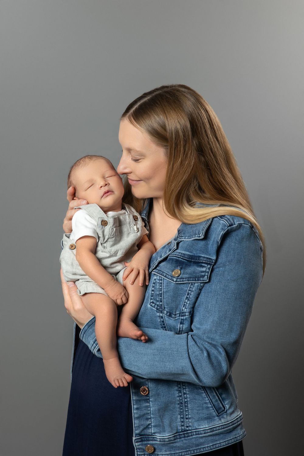 A young mother in a denim jacket tenderly holds and kisses her sleeping newborn baby against a gray background.