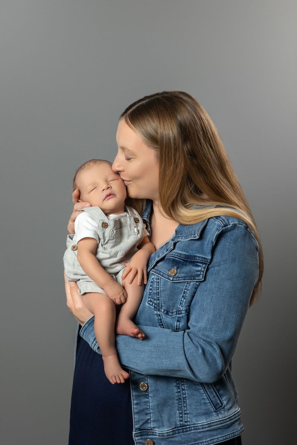A young mother in a denim jacket tenderly kisses her sleeping newborn baby against a gray background.