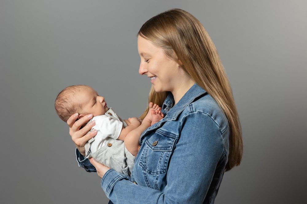 Young woman in denim jacket smiling while cradling a newborn baby against a gray studio background.