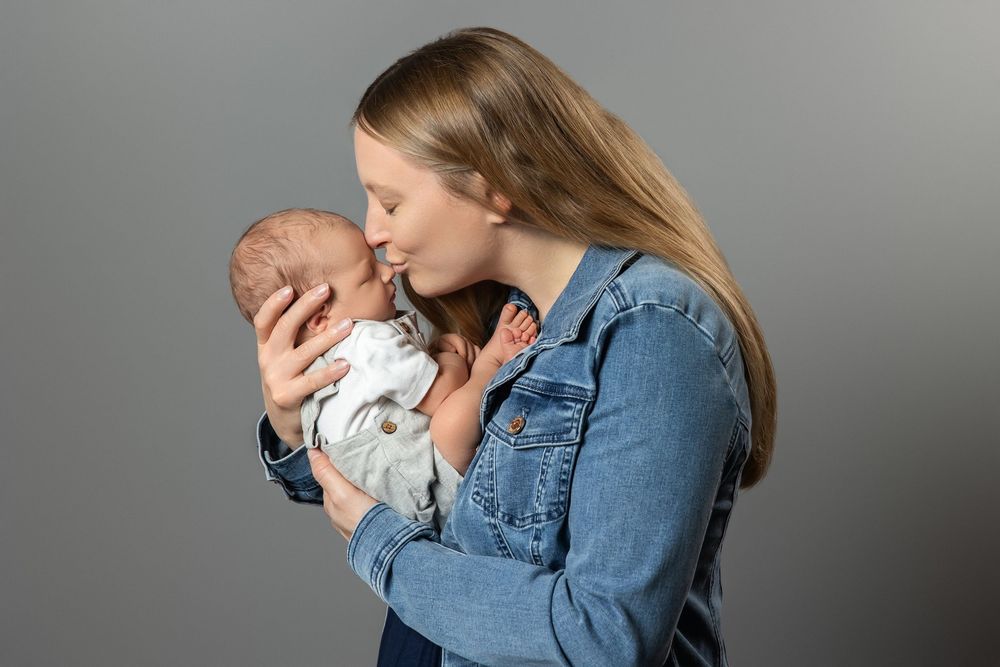 Young mother in denim jacket tenderly kissing her newborn baby against a gray studio background.