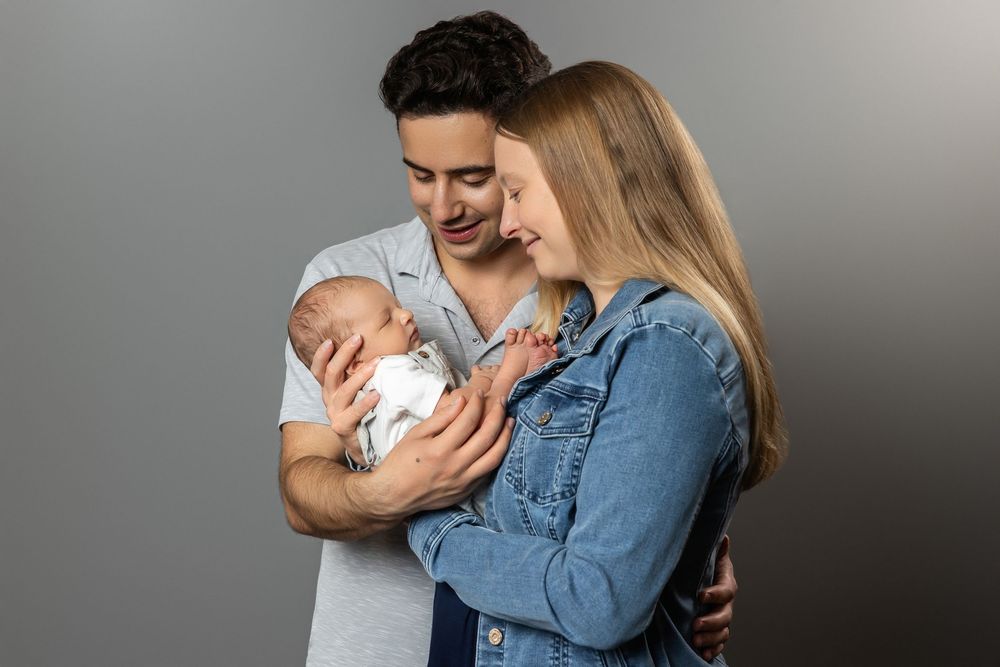 Young couple smiling and holding a sleeping newborn baby against a gray studio background.