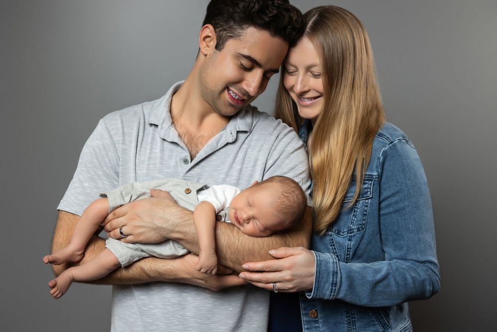 Happy couple smiling and holding their newborn baby together against a gray studio background.