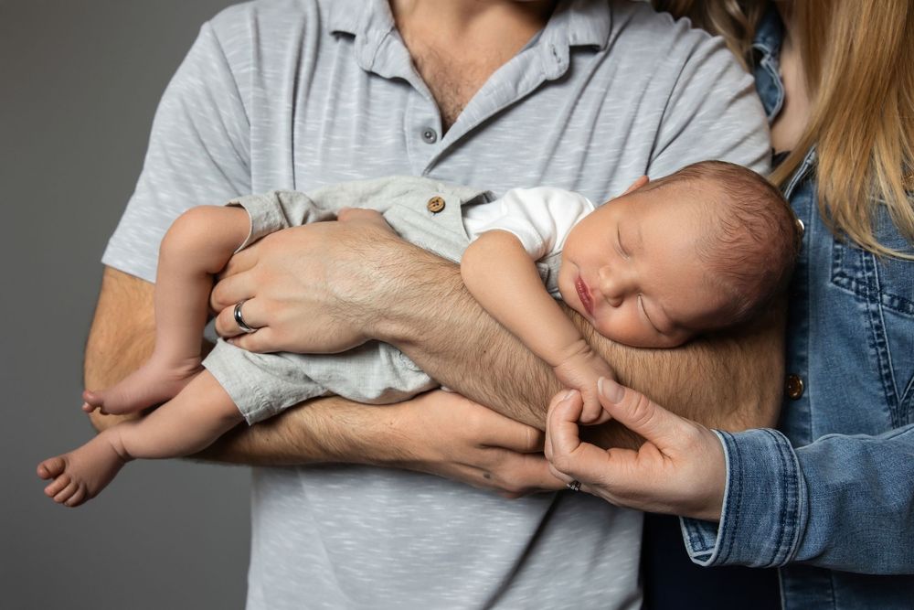 Parents cradling their sleeping newborn baby, with father holding infant securely in both hands against gray background.