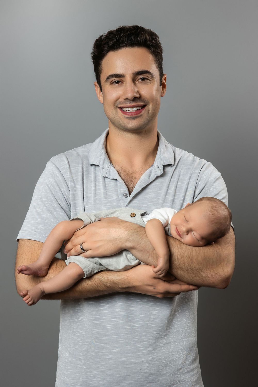 Smiling young father holding sleeping newborn baby in his arms against a gray background.