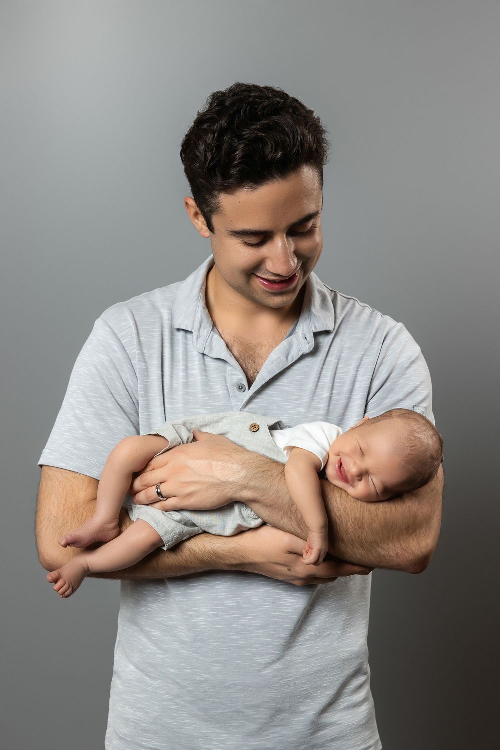 Smiling young father holding sleeping newborn baby in arms, wearing gray polo shirt against gray background.