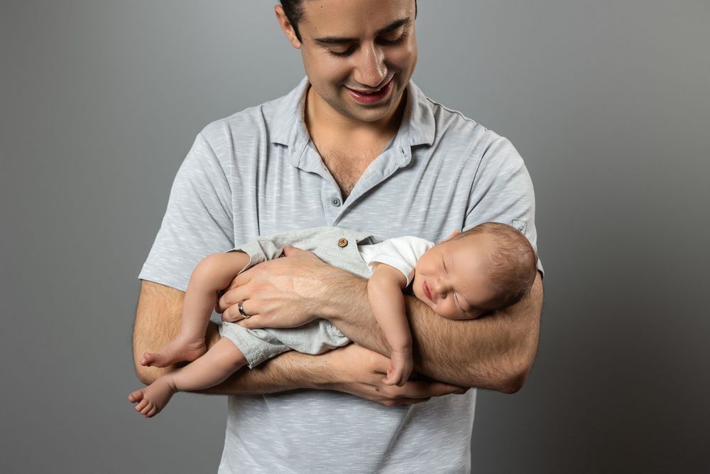 Smiling man in gray shirt holding two sleeping newborn babies in his arms against gray background.