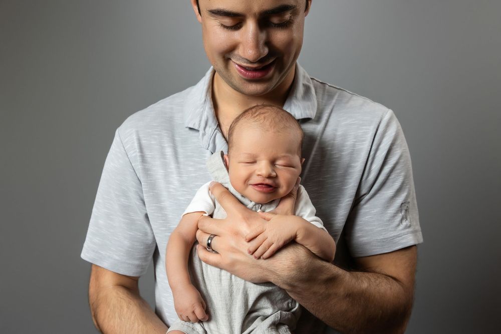 Smiling young father holding sleeping newborn baby against gray studio background.