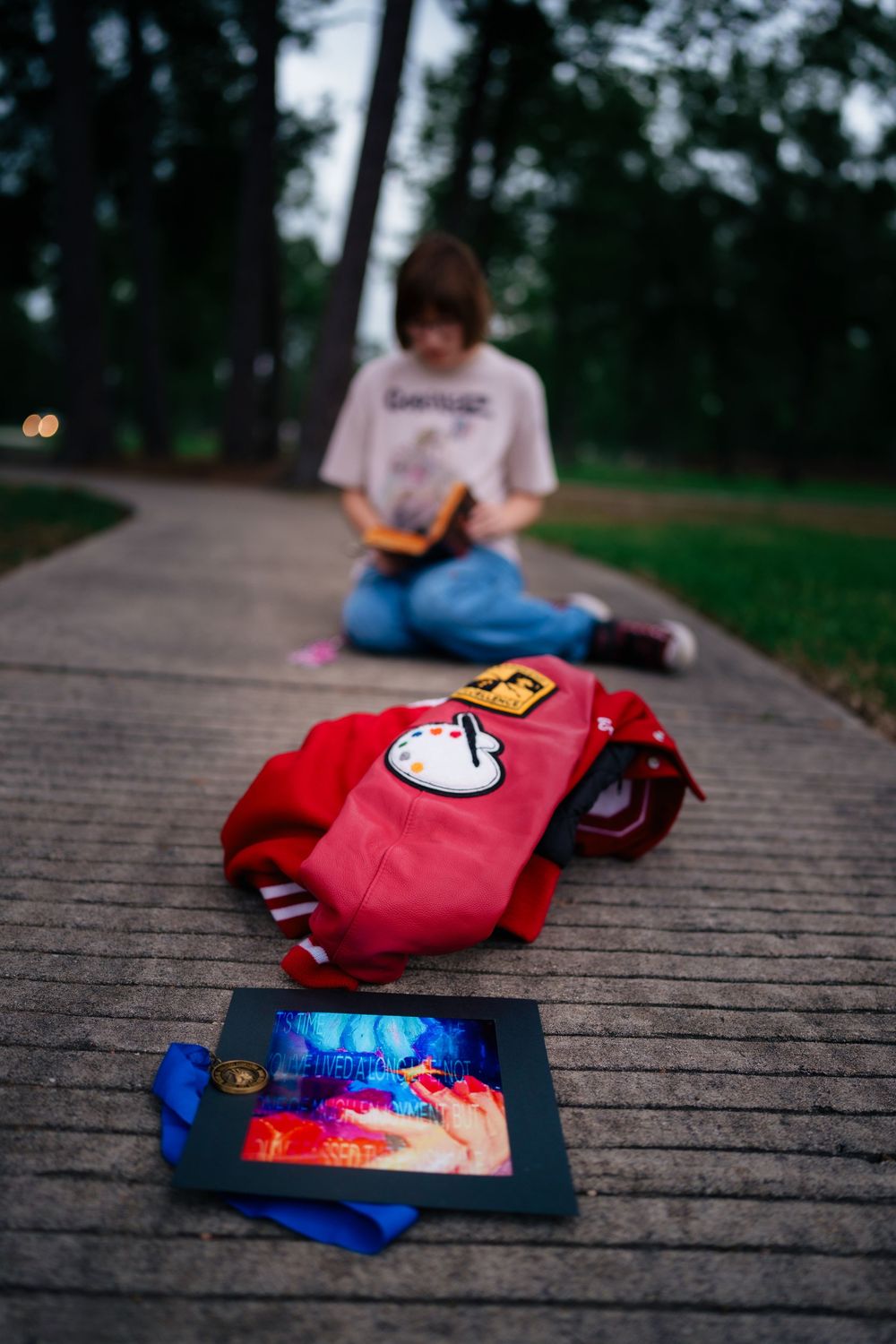 A red backpack with patches and colorful photo on a path, with a girl reading a book in the background.