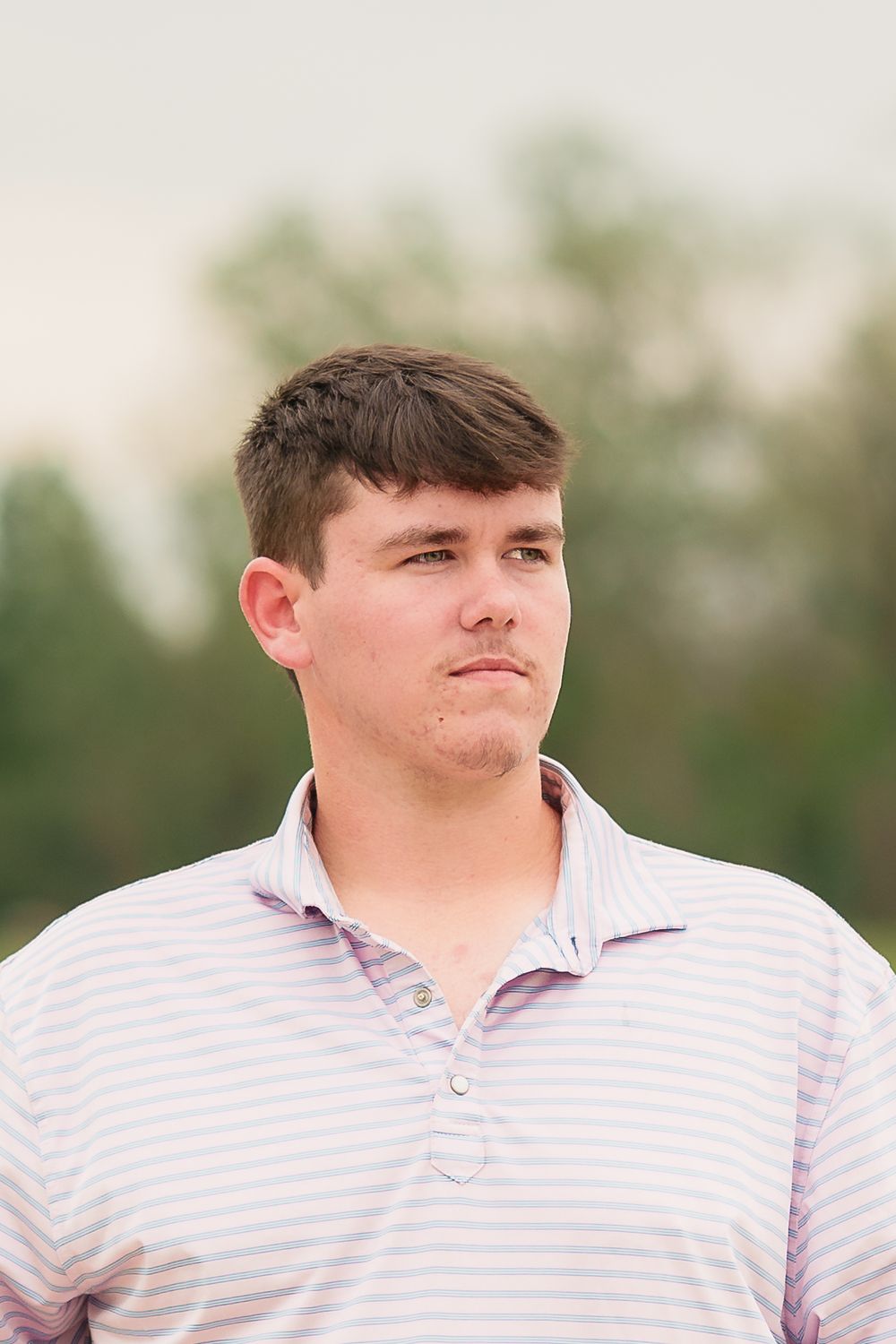 Young man in a pink striped polo shirt standing outdoors with green trees in the background.