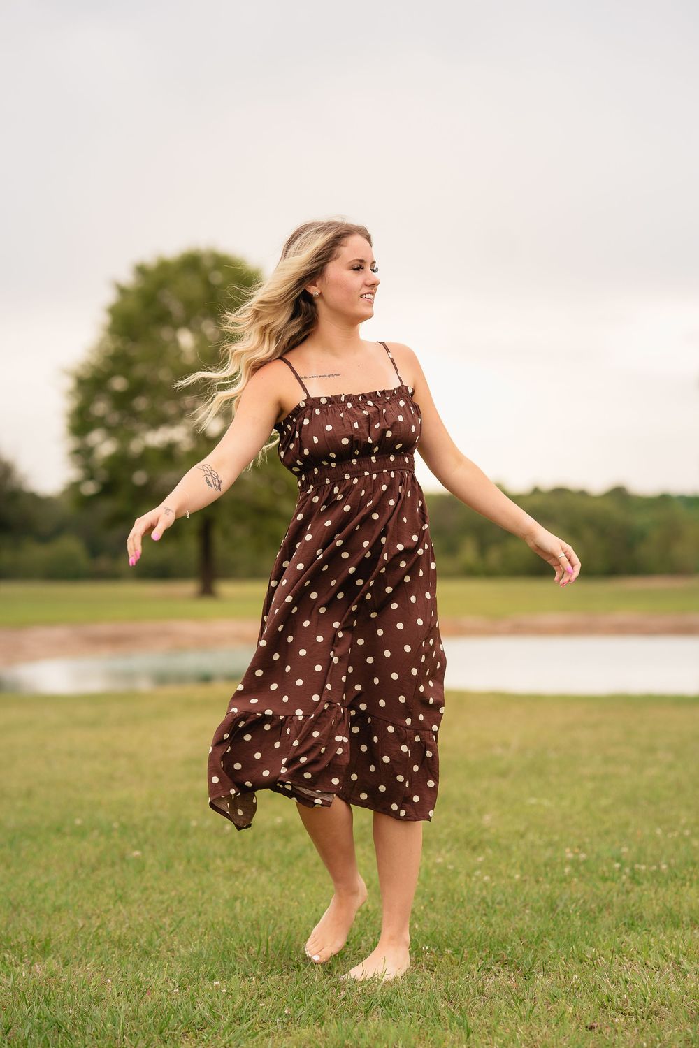 Blonde woman twirling in a brown polka dot sundress barefoot on green grass near a pond.
