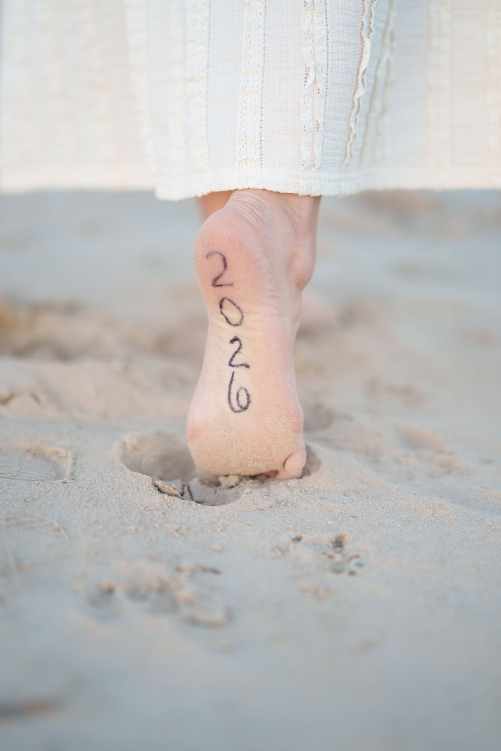 Close-up of a bare foot on sandy beach with '2026' written on the heel, wearing a white knit dress.