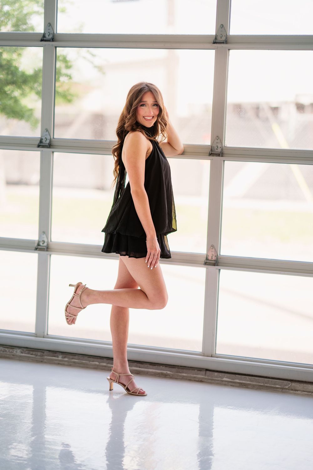 Young woman in black romper and nude heels posing confidently by large window with natural light.