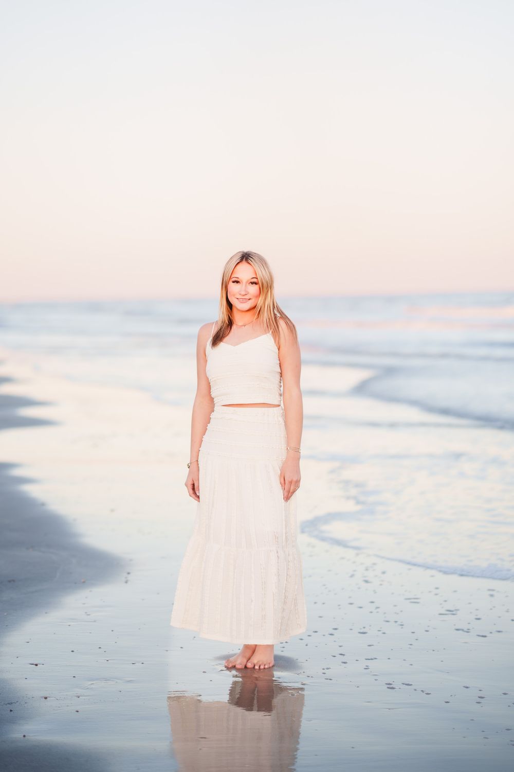 Blonde woman in white crop top and maxi skirt standing barefoot on a reflective beach at sunset.