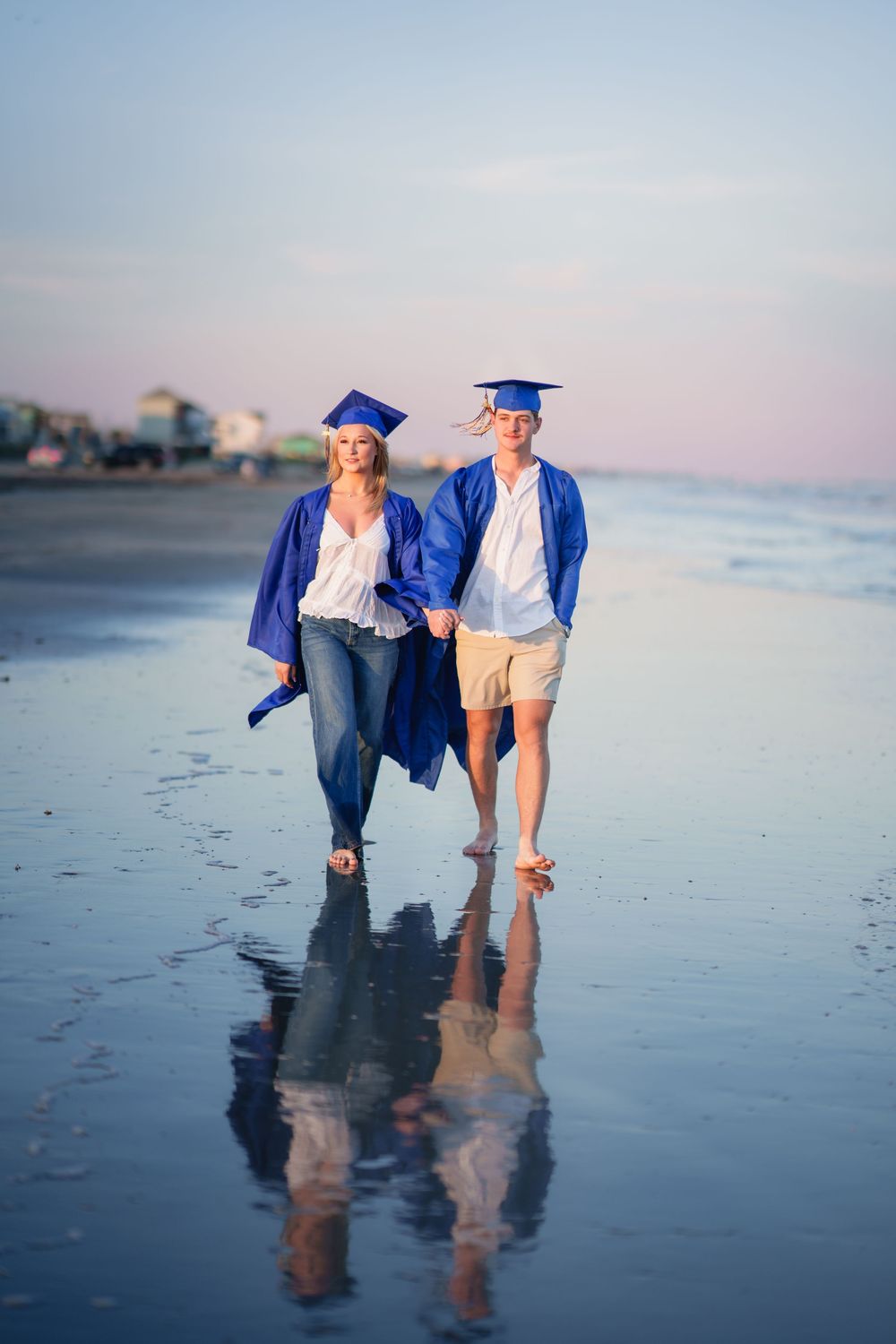 Two graduates in blue caps and gowns walk hand-in-hand on a reflective beach at sunset.