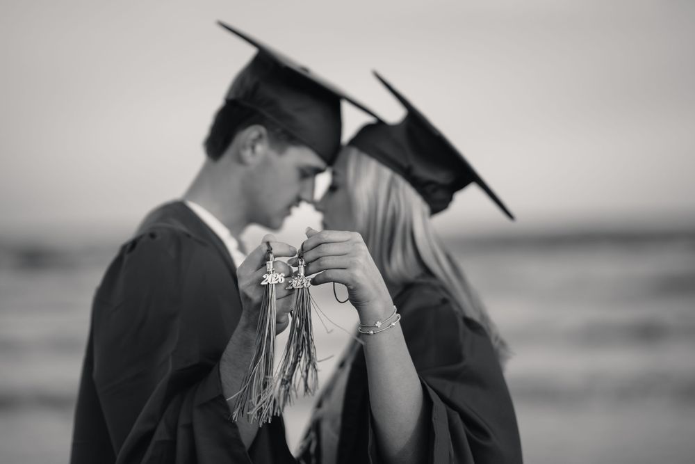 Two graduates in caps and gowns share a romantic moment on a beach in this black and white photo.