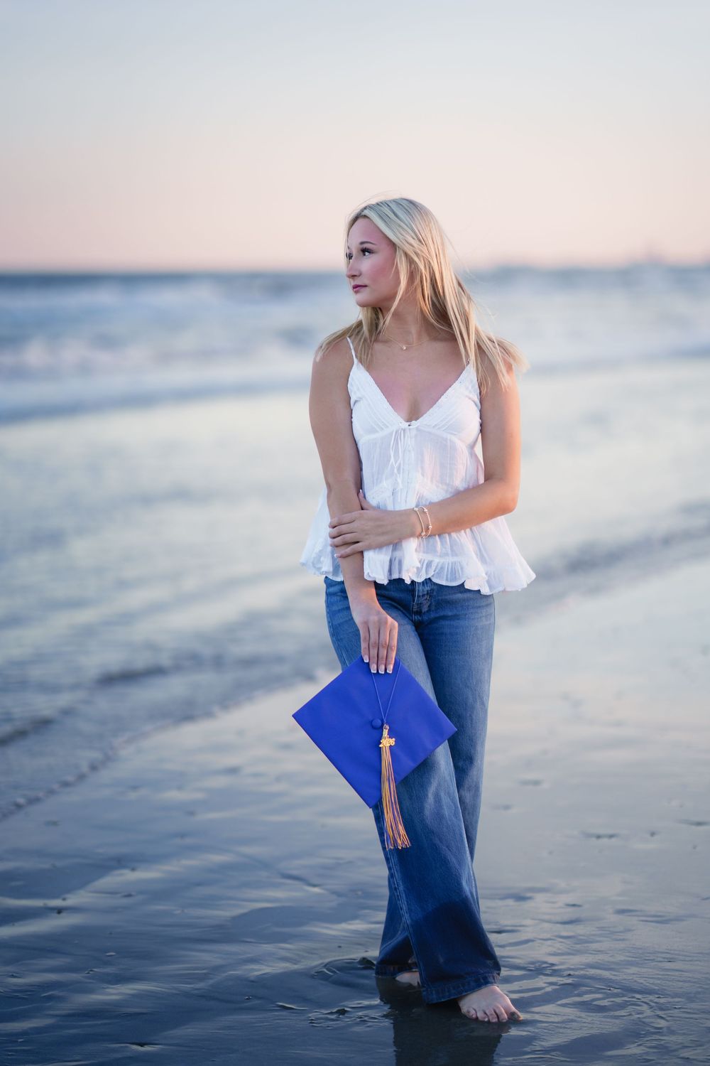 Blonde graduate holds blue cap on beach at sunset, wearing white top and jeans, standing in shallow water.