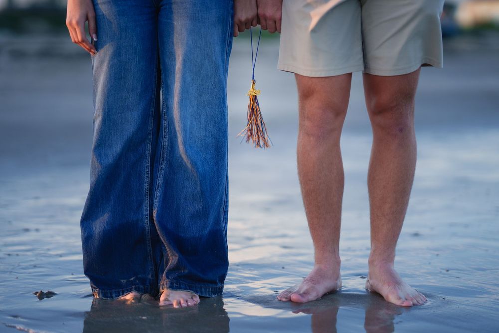 Two people standing barefoot on a wet beach, one in blue jeans, holding a decorative tassel together.
