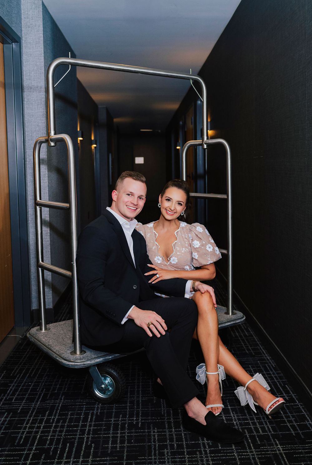 Couple posing on a hotel luggage cart in a modern hallway, man in black suit and woman in floral dress with heels.