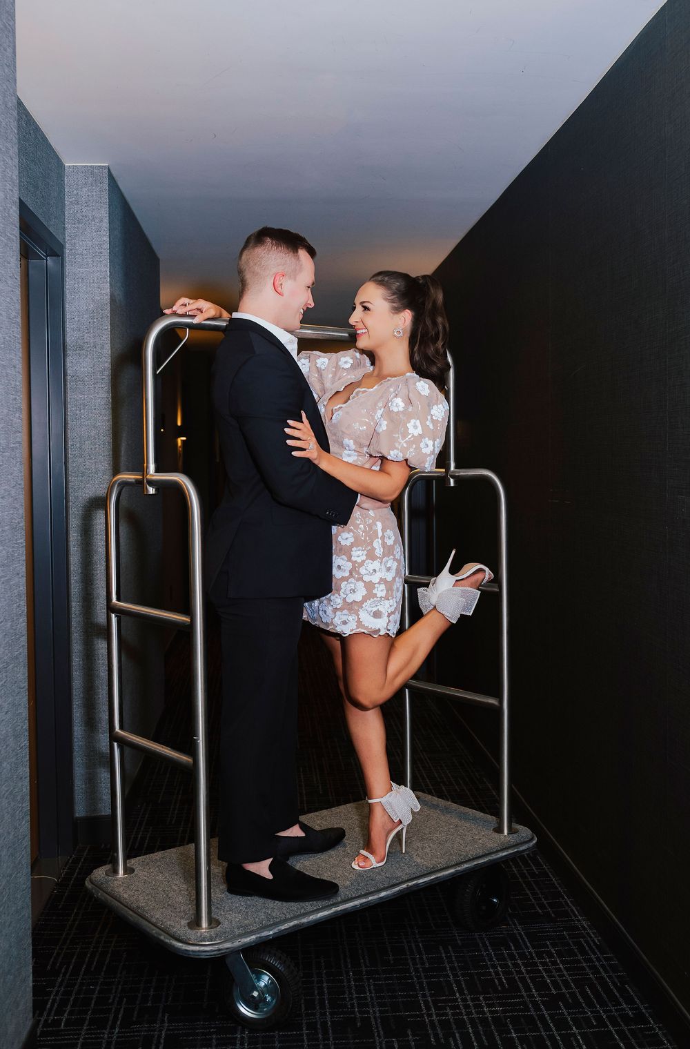 Couple posing romantically on a hotel luggage cart in a hallway, dressed in formal attire.