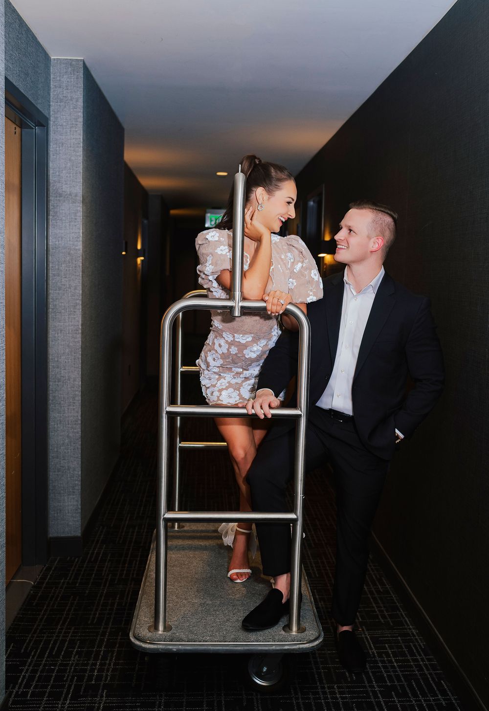 A couple poses playfully on a hotel luggage cart in a hallway, woman in floral dress, man in dark suit.