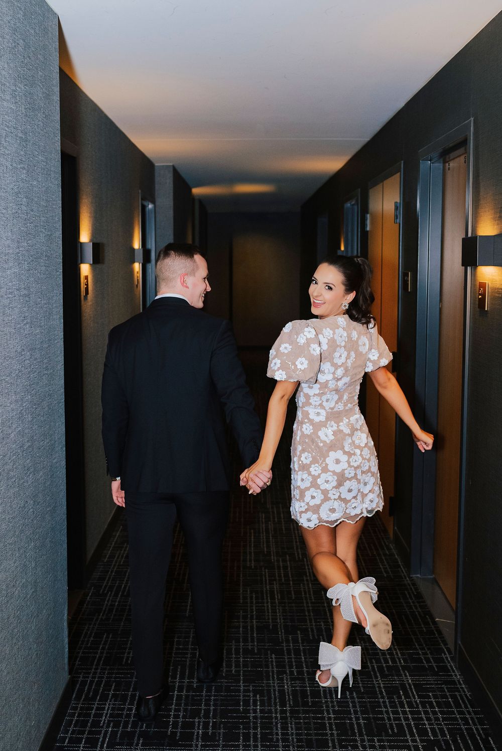 Couple holding hands walking down a modern hotel hallway, woman in floral mini dress and white heels, man in black suit.