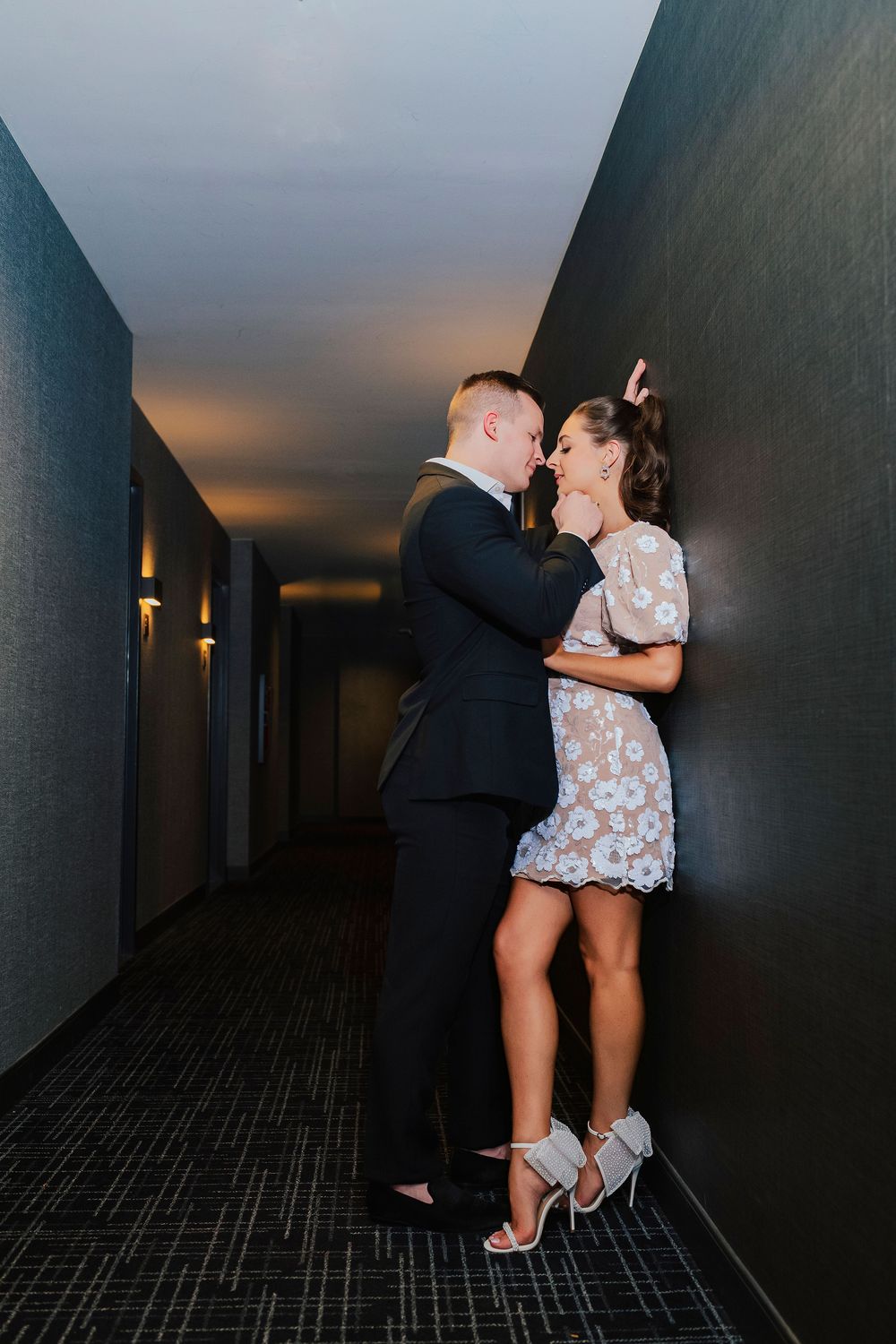 Couple in formal wear sharing a romantic moment in a dimly lit hotel corridor, man in suit, woman in lace mini dress.