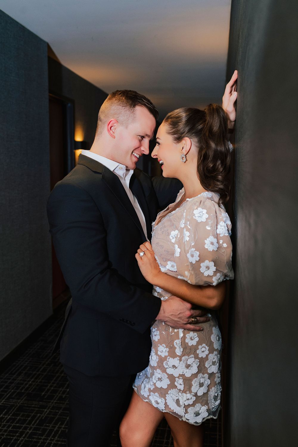 Couple in formal attire sharing a romantic moment, smiling at each other against a dark wall in dim lighting.