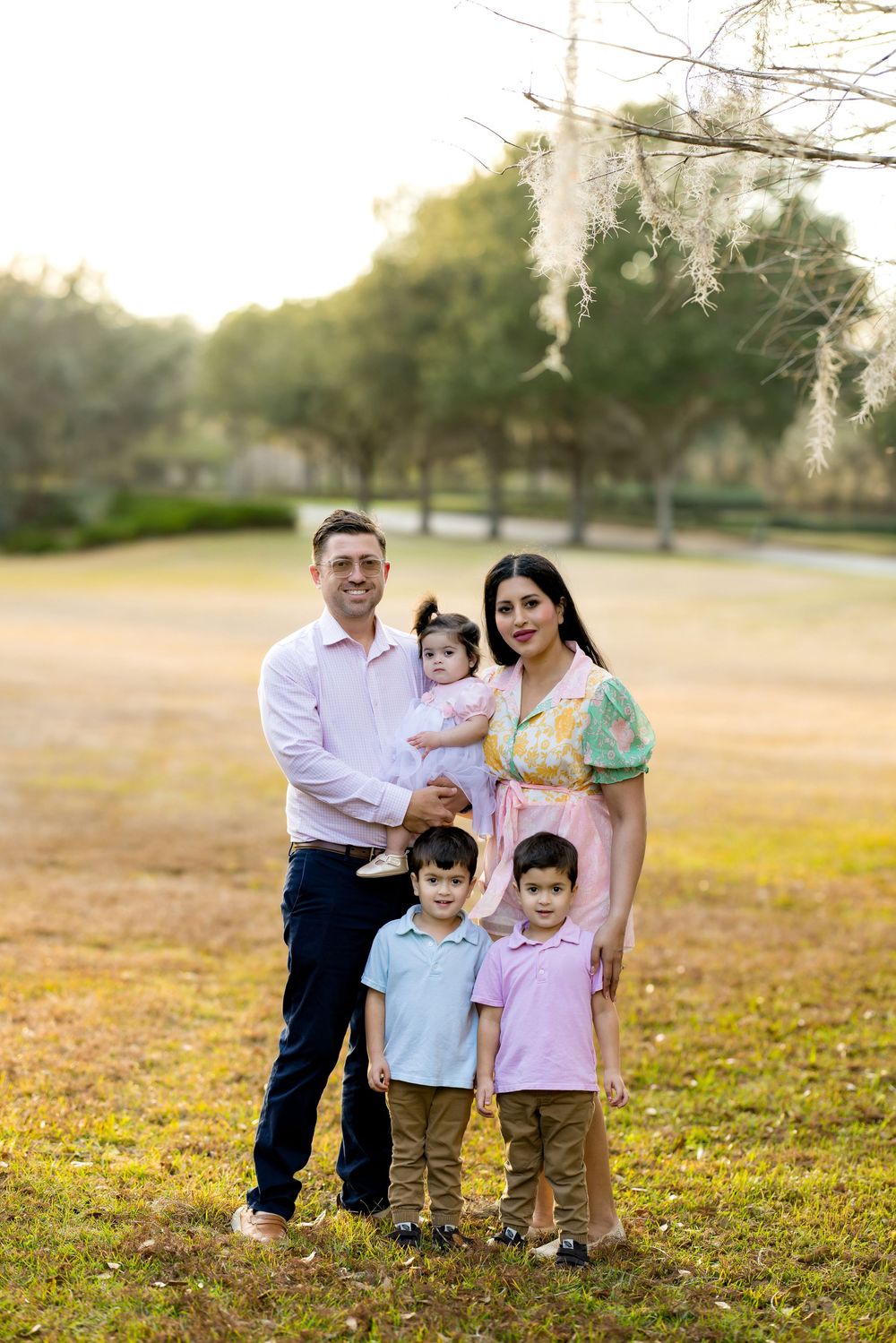 Family of five posing outdoors in a park during golden hour, parents with three young children smiling.