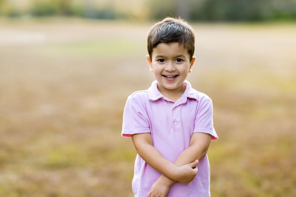 A smiling young boy in a pink polo shirt stands outdoors with arms crossed in a sunny field.