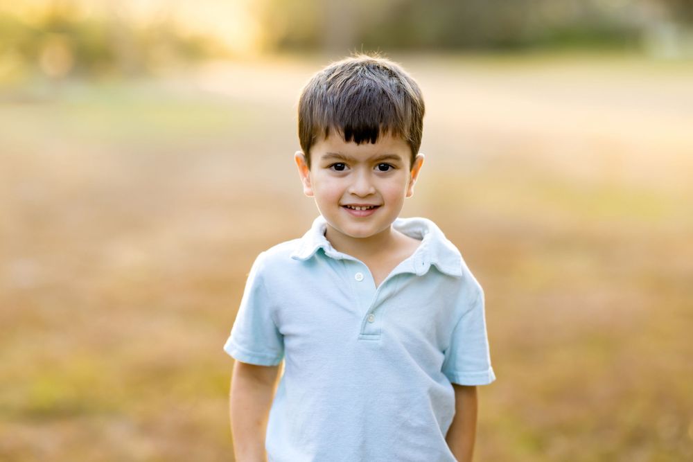 Smiling young boy in a light blue polo shirt standing outdoors in a sunny field with golden bokeh background.