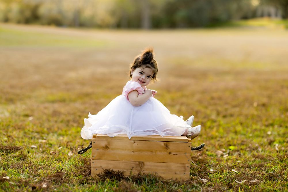 A cute toddler girl in a white dress sits in a wooden crate outdoors in a sunny green field.