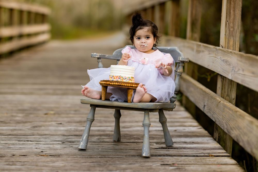 Toddler girl in pink dress sitting in vintage wooden chair on boardwalk with small birthday cake on stool.