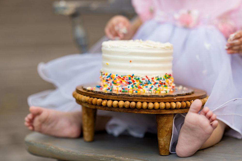 Baby's first birthday cake smash with white frosted cake topped with colorful sprinkles on wooden stand.