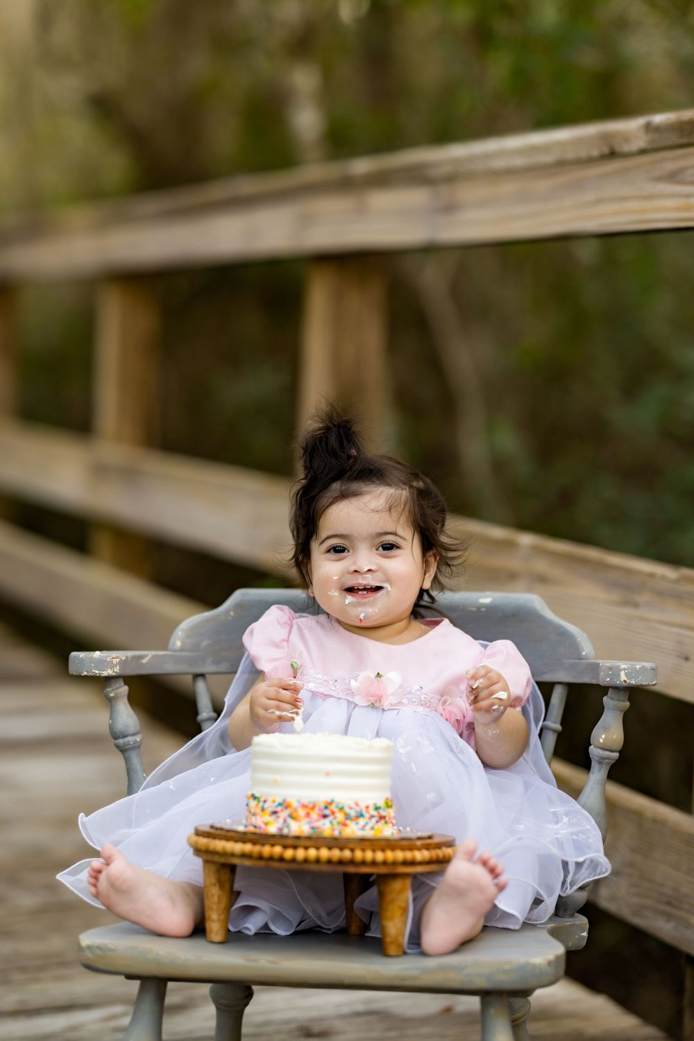 Baby girl in pink dress smiling at her first birthday cake smash on a wooden bridge outdoors.