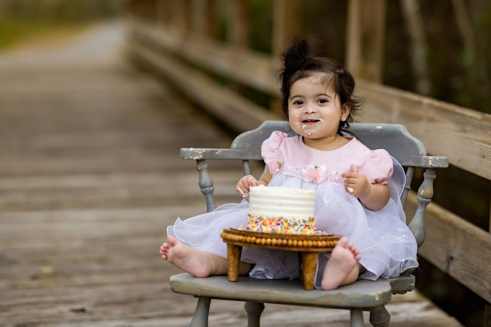 Toddler girl in pink dress sitting on chair with birthday smash cake on wooden bridge outdoor photo.