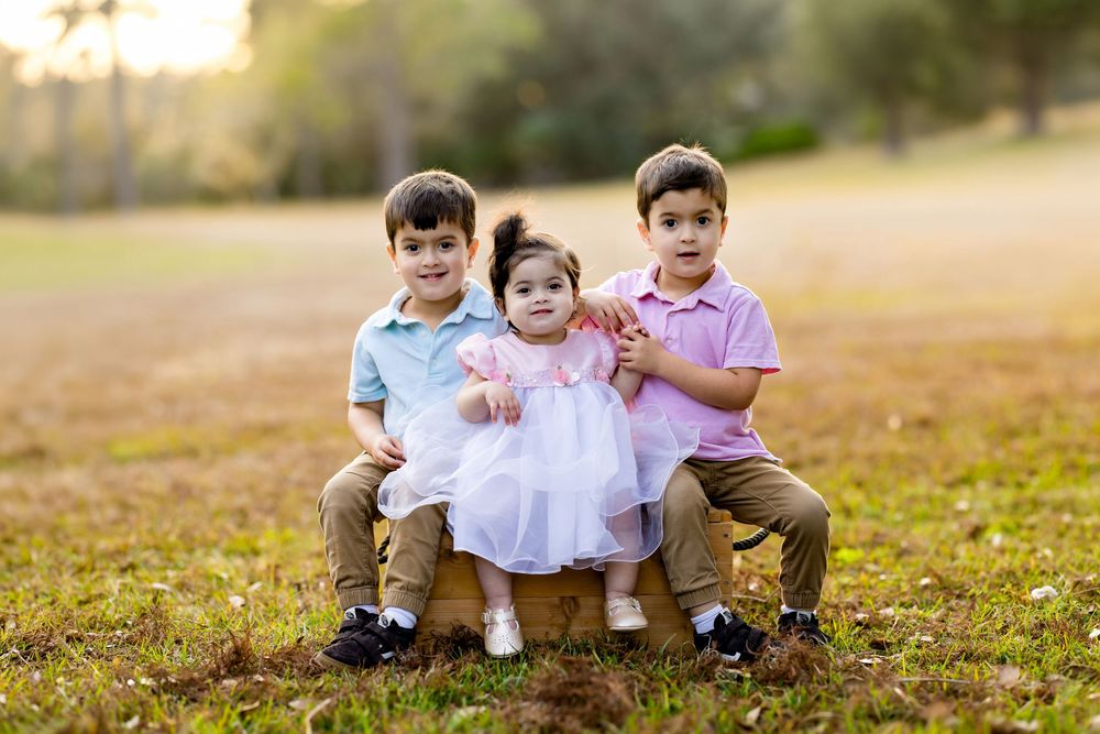 Three young children pose outdoors on a wooden box, two boys in pastel shirts and a baby girl in a white pink dress.