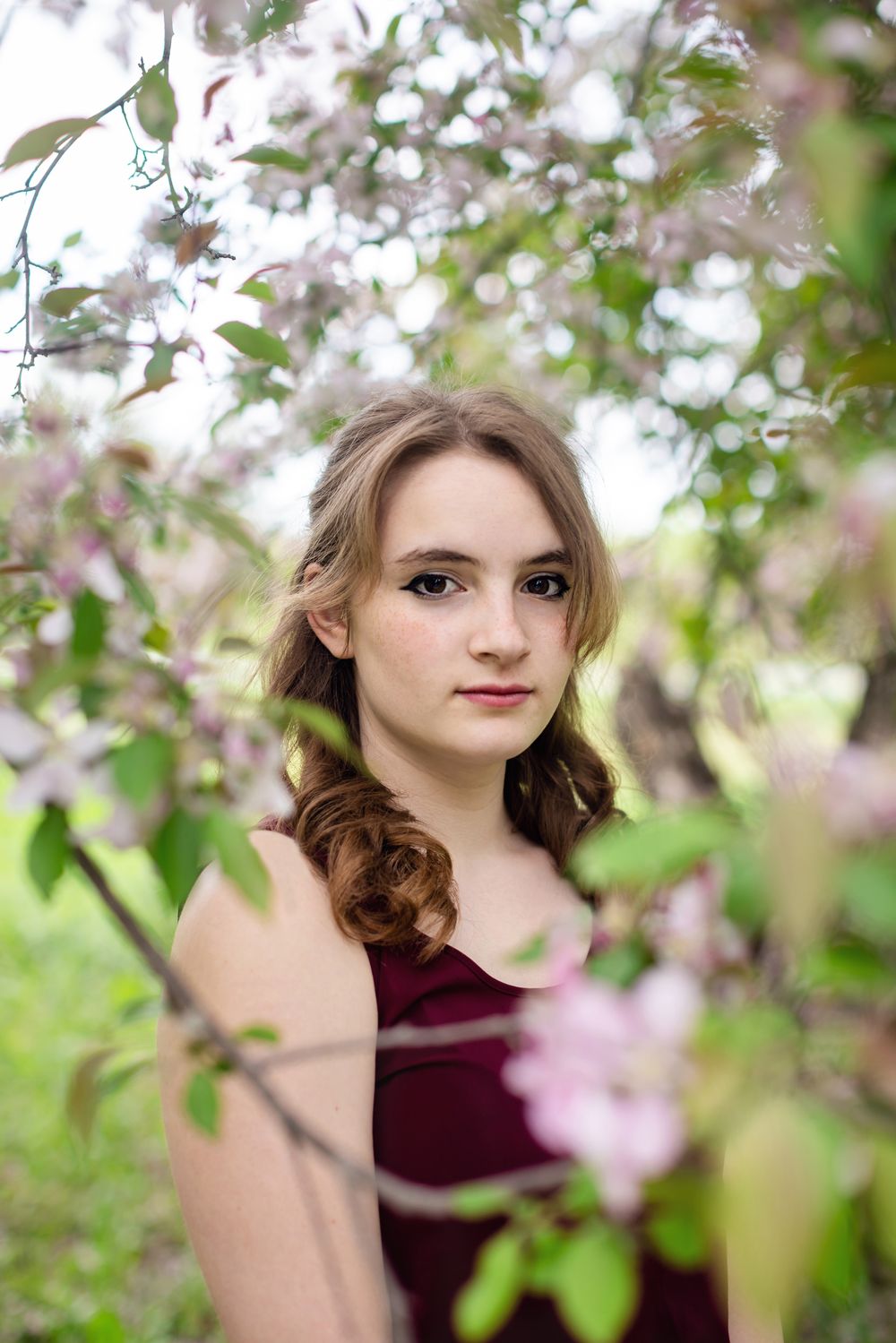 Young woman in burgundy dress surrounded by pink blossoming spring tree branches in a garden.