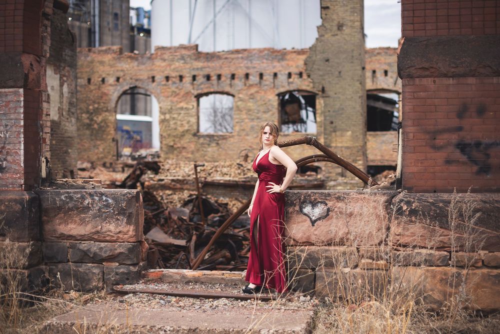 Woman in flowing red dress standing among abandoned brick ruins in an industrial wasteland setting.