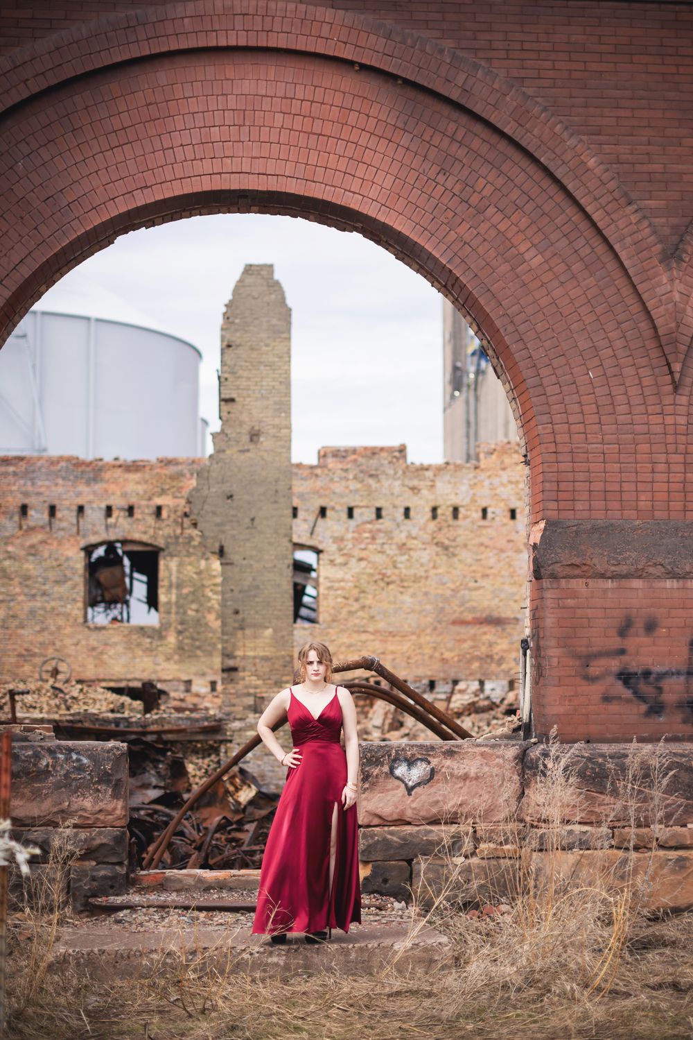 Woman in red dress standing under large brick arch at abandoned industrial building ruins.
